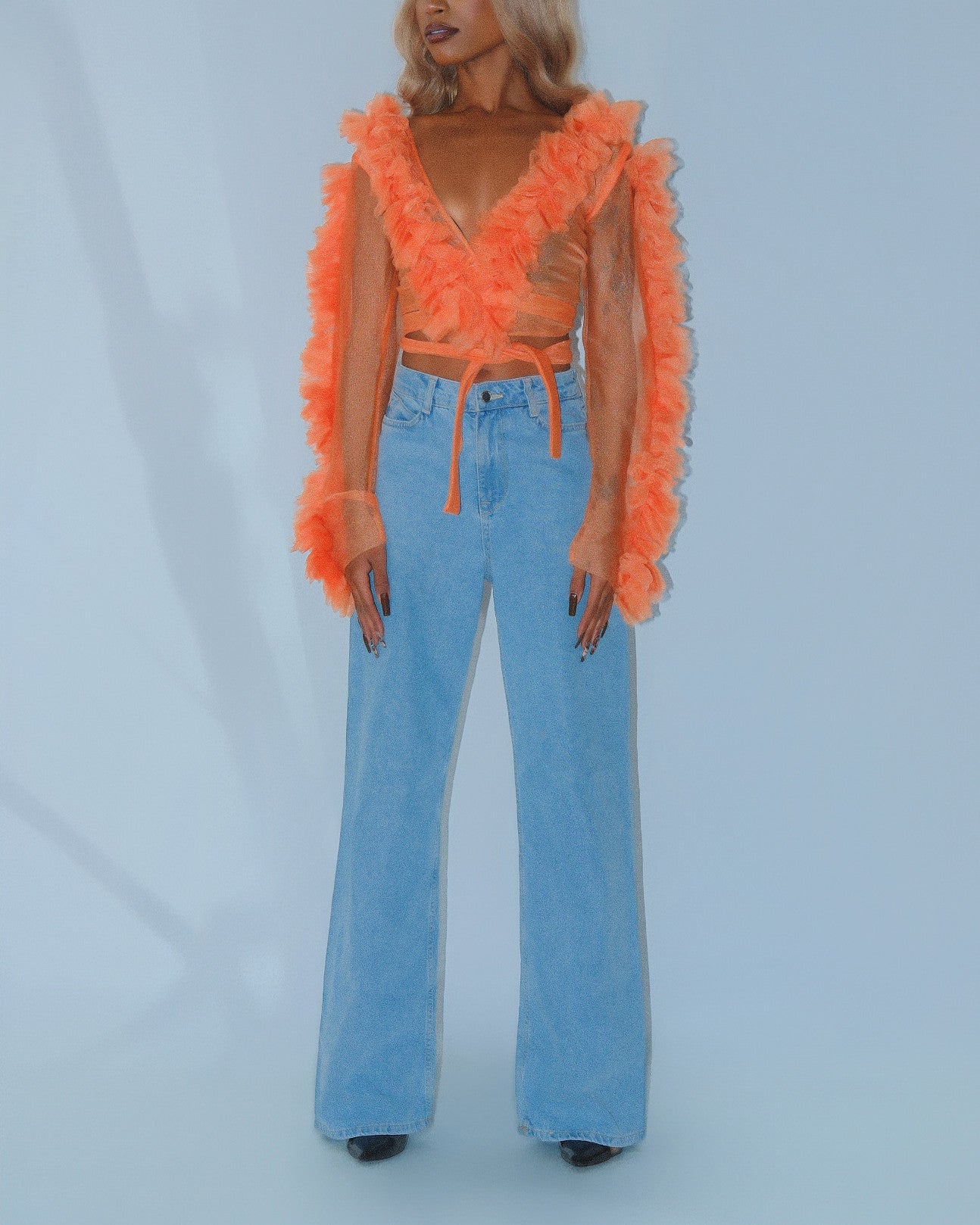 Woman wearing designer orange ruffled top and blue jeans against a light gray background. Dimaz